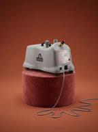 J-4000 Jiffy garment steamer with a black head, silver metal rod, light gray control base, and black cord, displayed on a round red pedestal against a warm rust-orange studio background - Sold by: Best Steamers