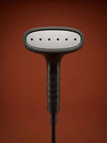 J-4000 Jiffy garment steamer with a black head, silver metal rod, light gray control base, and black cord, displayed on a round red pedestal against a warm rust-orange studio background - Sold by: Best Steamers