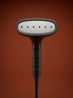 J-4000 Jiffy garment steamer with a black head, silver metal rod, light gray control base, and black cord, displayed on a round red pedestal against a warm rust-orange studio background - Sold by: Best Steamers