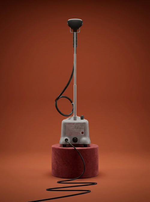 J-4000 Jiffy garment steamer with a black head, silver metal rod, light gray control base, and black cord, displayed on a round red pedestal against a warm rust-orange studio background - Sold by: Best Steamers